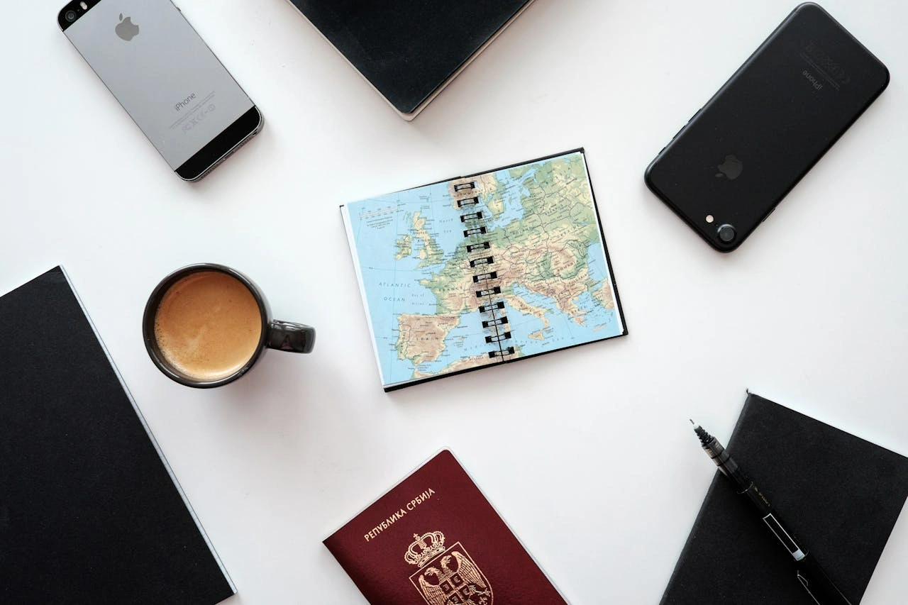 white desk with travel essentials like passport, maps, notebook, and phone placed on top