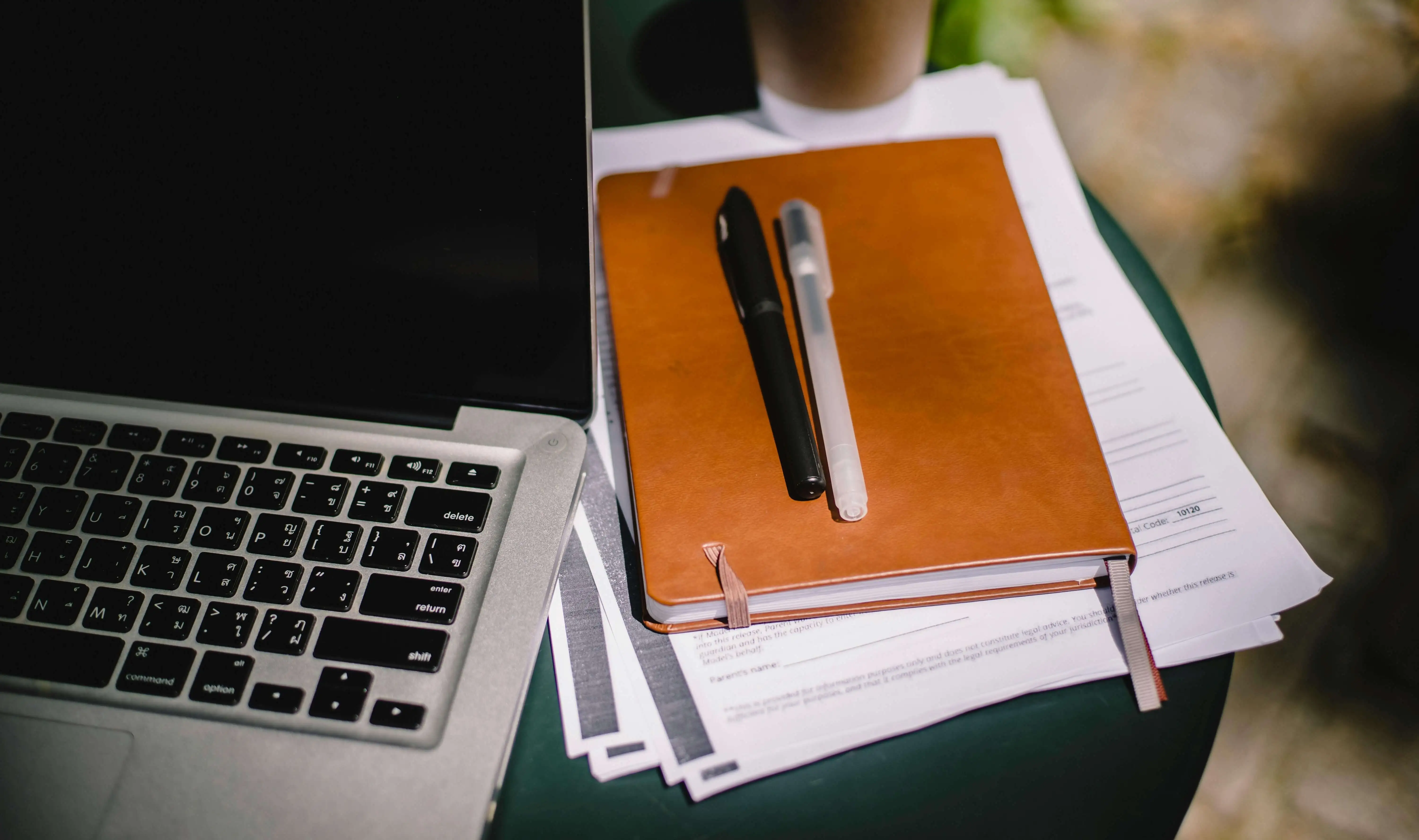 Laptop next to a notebook and some loose papers and pens on a small desk